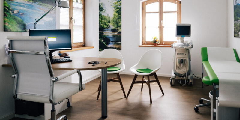 Praxisfotos Dr. E. Seidel - Juli 2026-146 Helles Behandlungszimmer mit einem Tisch für die Patientenbetreuung, einer Untersuchungsliege und medizinischen Messgeräten